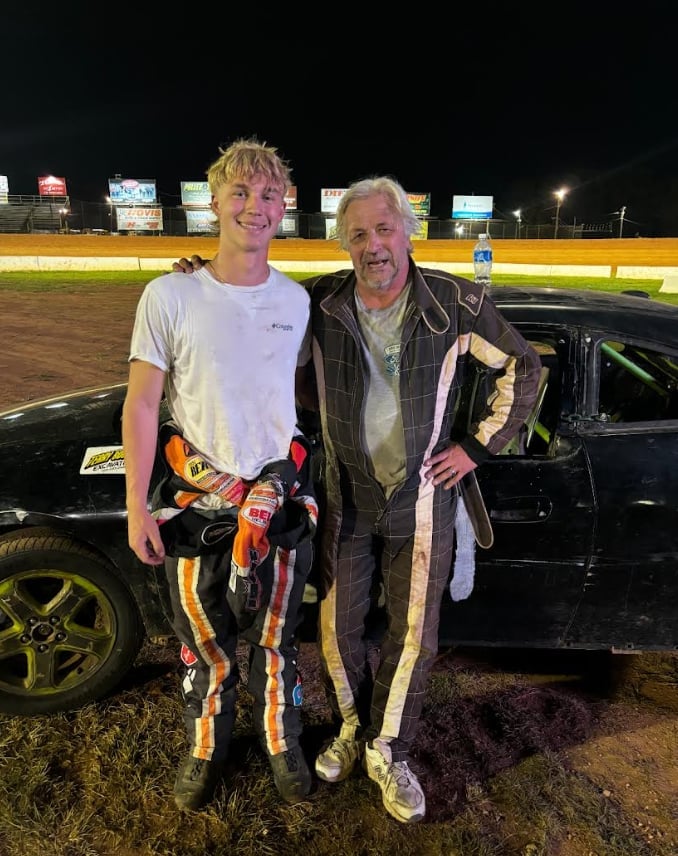 Two men posing at a race track at night, holding a toy car, standing next to a black vehicle