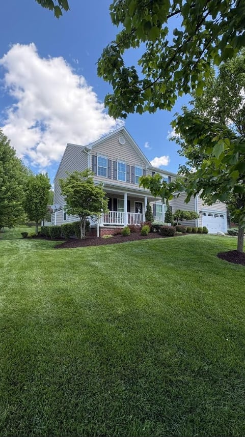 Two-story white house with black shutters, front porch, and attached garage on manicured lawn surrounded by green trees
