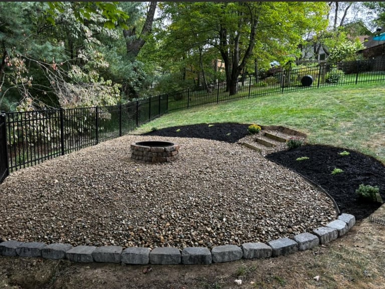 Backyard fire pit area with gravel, stone border, and black mulch landscaping surrounded by green lawn and trees