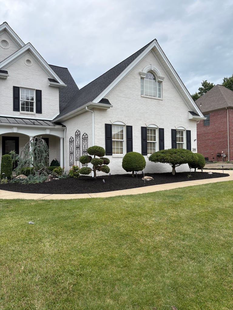 White brick two-story house with black shutters, manicured landscaping, and lawn under cloudy sky