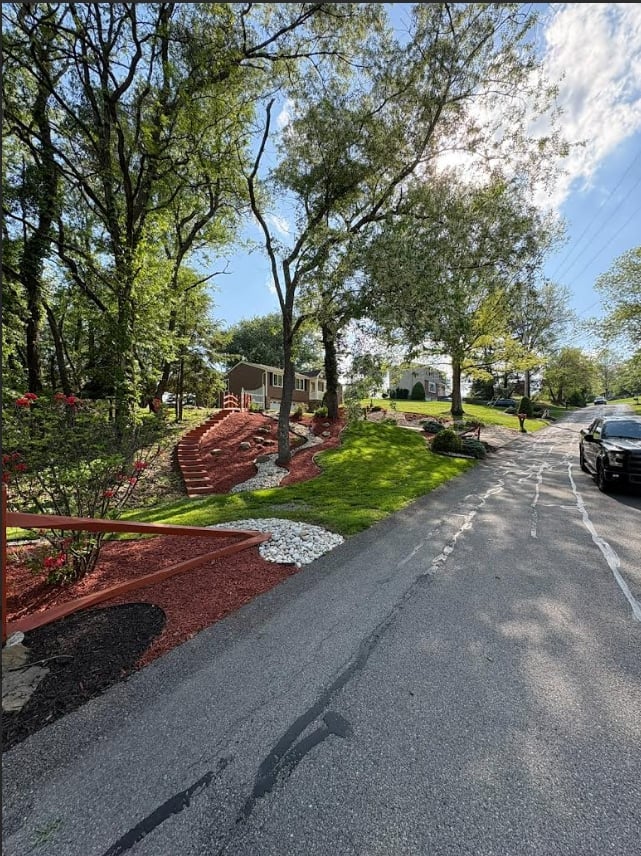 Residential driveway lined with mature trees, red mulch landscaping, playground equipment, and a parked car on a sunny day