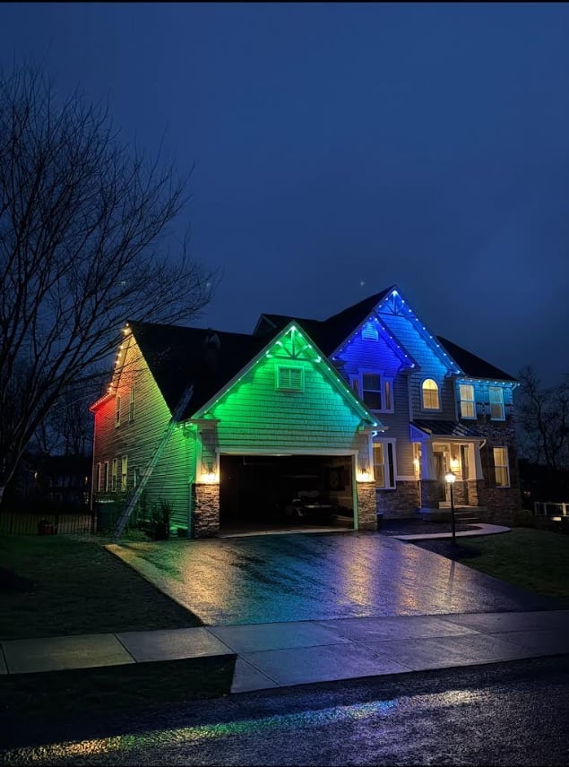 Two-story house illuminated with colorful LED lights in red, green, and blue at dusk, featuring a garage and bare trees