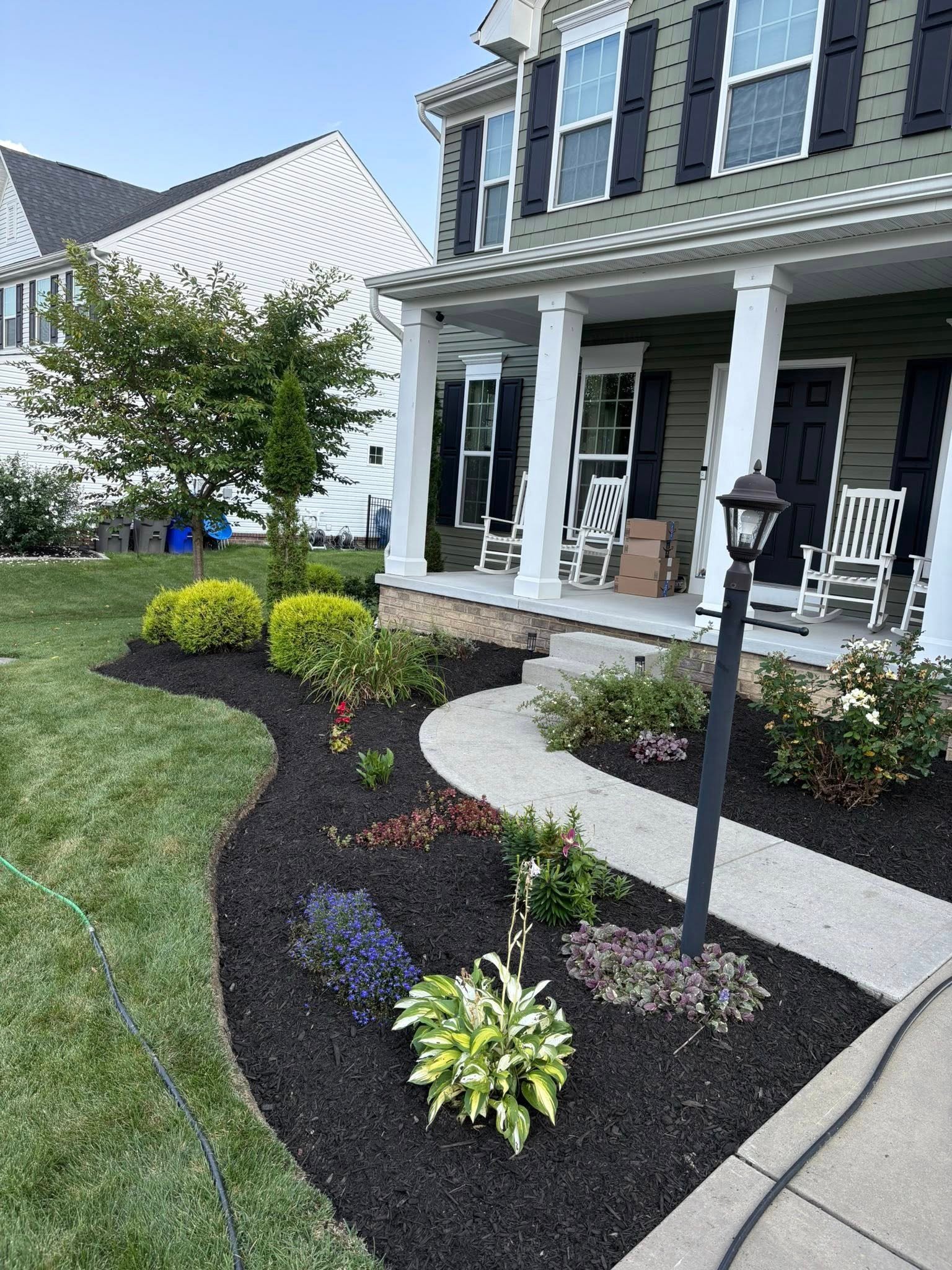 Green two-story house with white porch columns and manicured landscaping featuring flowering plants and shrubs in front