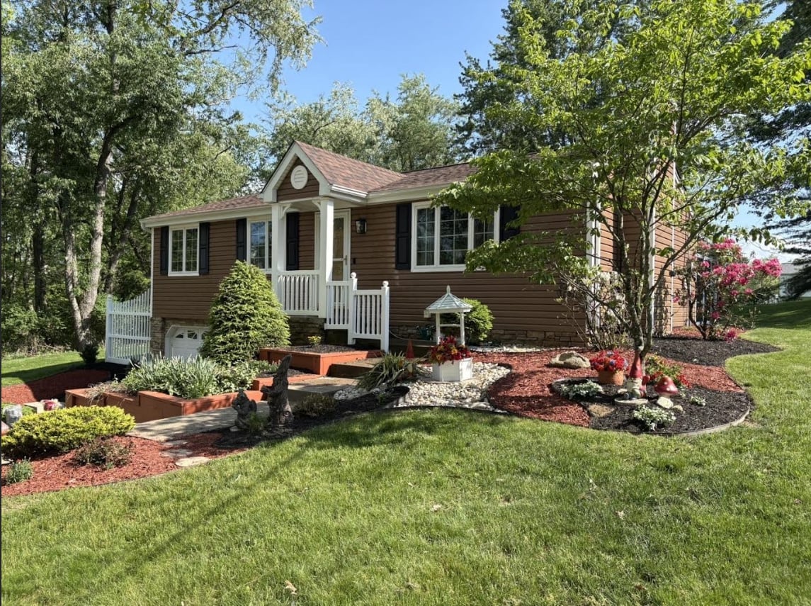 Brown single-story house with white columns and porch, surrounded by landscaped gardens with red mulch and flowering shrubs on a sunny day