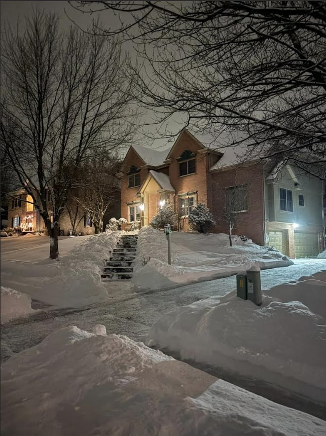 Brick house with peaked roof illuminated at night during winter, surrounded by snow-covered yard and bare trees