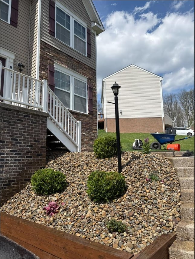 Residential yard with brick house, white deck, landscaped gravel bed with shrubs and lamp post, neighboring white garage visible