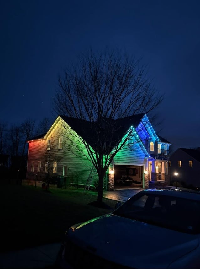 House lit with colorful green and blue exterior lights at dusk, bare tree silhouetted in front, driveway with parked cars