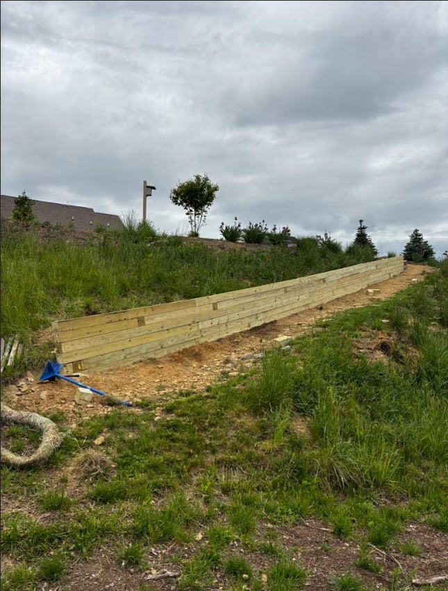 Newly constructed wooden retaining wall on hillside with green grass, overcast sky, and trees in background