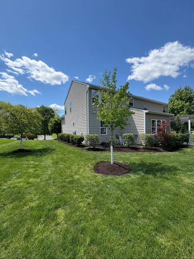 Two-story gray vinyl house with green lawn, young tree, landscaping, and clear blue sky with white clouds