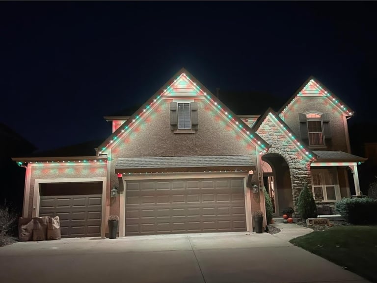 Two-story stone house at night decorated with green and pink holiday lights outlining the roofline and gables